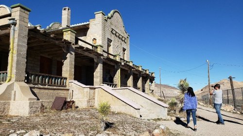 Two people walk towards an old, abandoned building under a clear blue sky. Desert landscape surrounds the area.