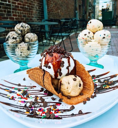 A dessert plate featuring a waffle cone filled with ice cream, chocolate sauce, sprinkles, and two bowls of ice cream.