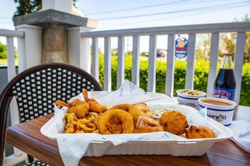 A seafood platter with fried shrimp, onion rings, and hush puppies, accompanied by a soda, on a table outdoors.