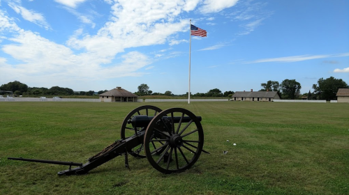 A historic cannon on green grass with an American flag waving in the background and buildings in the distance.