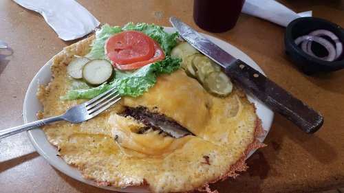 A plate with a large cheeseburger wrapped in an omelet, garnished with lettuce, tomato, pickles, and a knife.