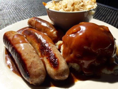 A plate with three grilled sausages, mashed potatoes topped with brown gravy, and a bowl of coleslaw.