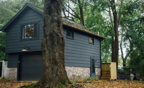 A dark gray house with a stone foundation, surrounded by trees and fallen leaves, featuring a garage and wooden steps.