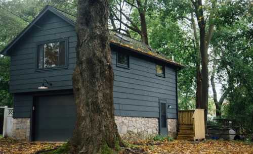 A dark gray house with a stone foundation, surrounded by trees and fallen leaves, featuring a garage and wooden steps.