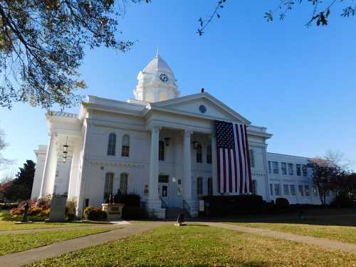 A white historic courthouse with a large American flag hanging from the front, surrounded by trees and clear blue sky.