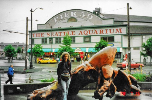 A person stands by a large sculpture in front of the Seattle Aquarium at Pier 59 on a rainy day.
