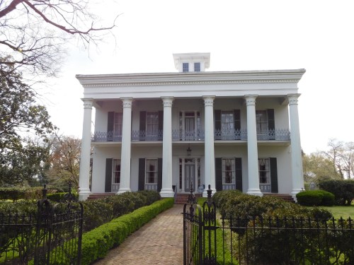 A grand white mansion with columns, surrounded by greenery and a wrought-iron fence, set against a cloudy sky.