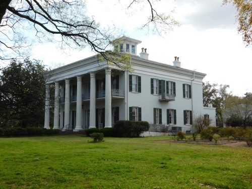 A large white mansion with columns, green shutters, and a manicured lawn under a partly cloudy sky.