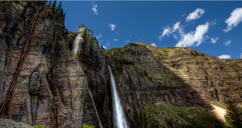 A scenic view of a waterfall cascading down rocky cliffs, with a house perched on the edge under a blue sky.