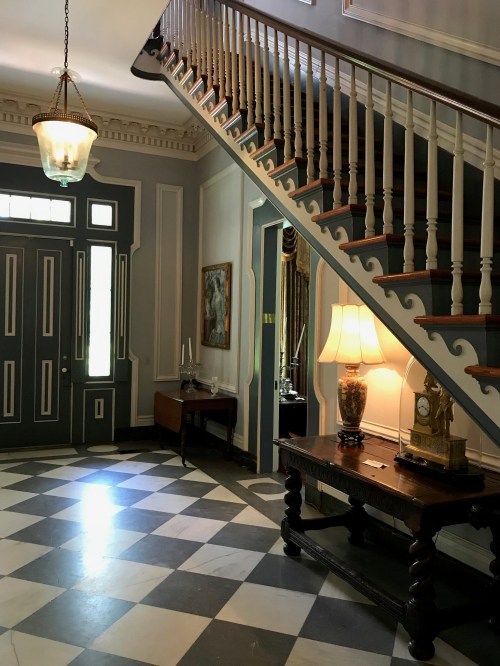 A spacious foyer with a staircase, checkered floor, vintage lamp, and decorative wall art.