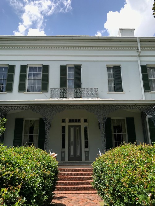 A historic house with green shutters, a balcony, and lush greenery in front, under a blue sky with clouds.