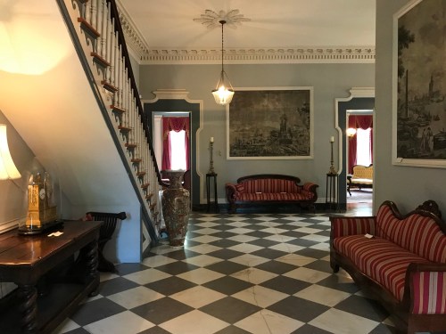 A spacious foyer with a checkered floor, staircase, vintage furniture, and large paintings on the walls.