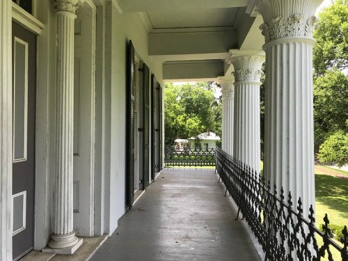 A covered porch with white columns and a decorative railing, overlooking a green lawn and trees.