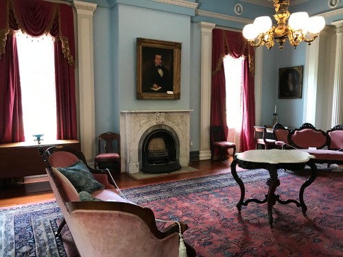 A historic room featuring elegant furniture, a fireplace, and decorative curtains, with portraits on the walls.