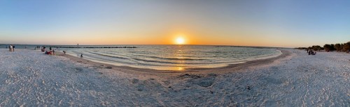 A panoramic view of a beach at sunset, with people walking along the shore and gentle waves lapping at the sand.