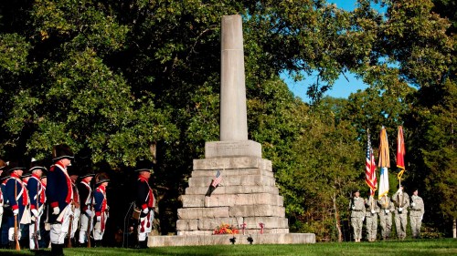 A monument surrounded by soldiers in uniform, with flags displayed, under a clear blue sky and trees in the background.