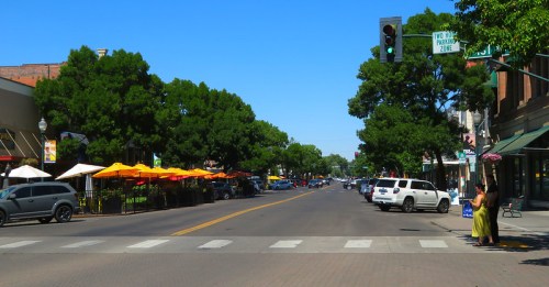 A sunny street scene with parked cars, outdoor dining areas, and green trees lining both sides of the road.