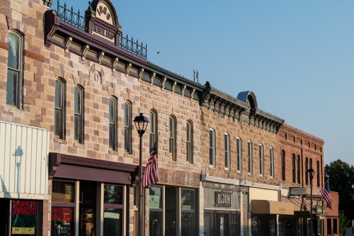 Historic brick buildings line a street, featuring shops and American flags under a clear blue sky.