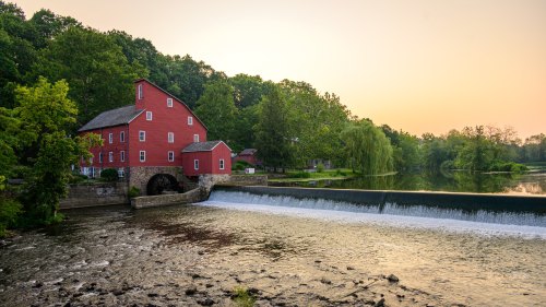 A red mill building beside a river, with a small waterfall and lush greenery in the background during sunset.