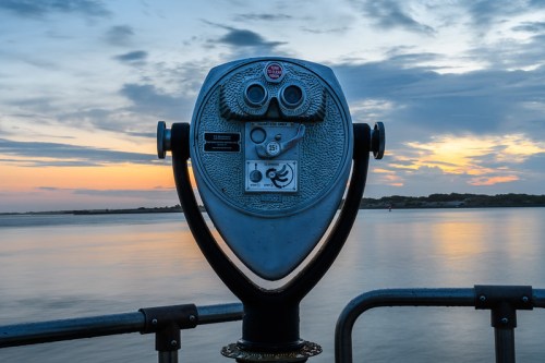 A coin-operated binocular viewer facing a calm body of water at sunset, with clouds in the sky.
