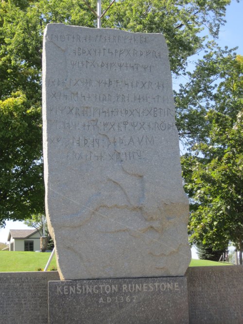 A large stone monument with ancient runes inscribed, labeled "Kensington Runestone A.D. 1362." Surrounded by greenery.