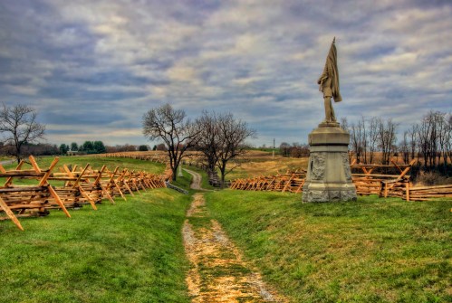 A statue stands at the end of a grassy path, surrounded by wooden fences and bare trees under a cloudy sky.
