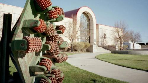 A close-up of a green and red agricultural tool in front of a modern building with a curved entrance and landscaped lawn.