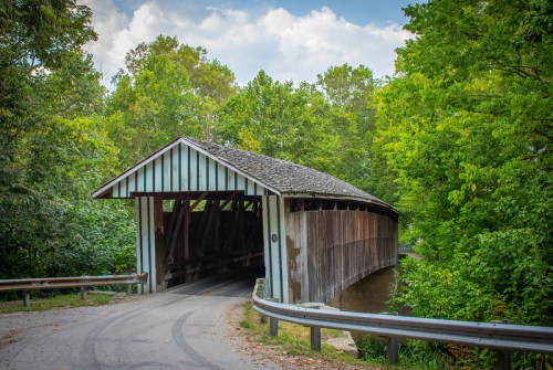 A rustic covered bridge surrounded by lush green trees, with a winding road leading to it.