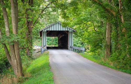 A covered bridge surrounded by lush green trees, leading to a quiet road.