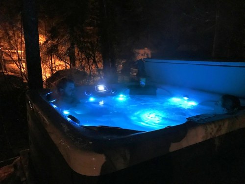 Two people relax in a hot tub at night, surrounded by trees and illuminated by blue lights.
