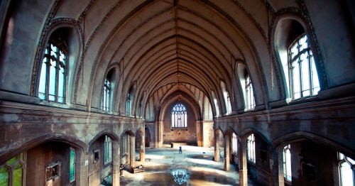 A spacious, abandoned hall with high arched ceilings and large windows, illuminated by soft natural light.