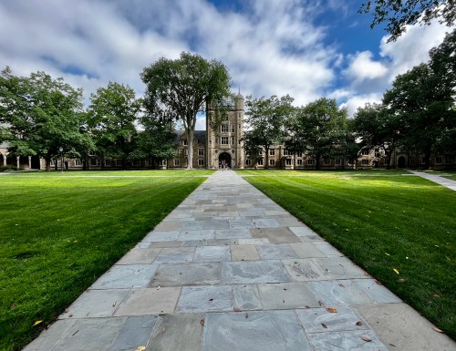 A stone pathway leads to a grand building surrounded by lush green trees and grass under a partly cloudy sky.