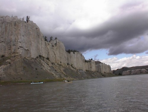 A river flows past towering cliffs under a cloudy sky, with a small boat visible near the shore.