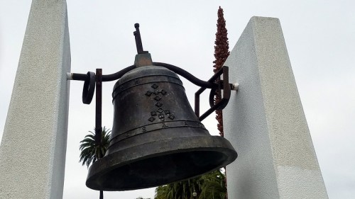 A large bronze bell suspended between two white pillars, surrounded by palm trees and a cloudy sky.
