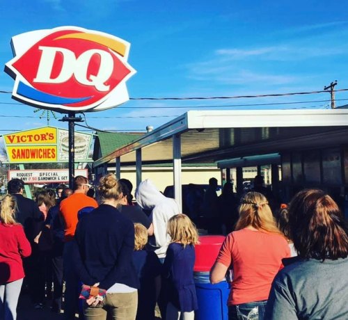 A crowd of people waiting in line outside a Dairy Queen and Victor's Sandwiches on a sunny day.