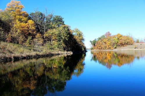 A serene lake surrounded by trees with autumn foliage, reflecting the clear blue sky and colorful leaves.