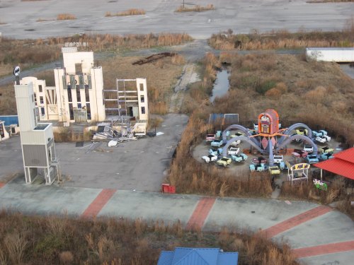 Abandoned amusement park with a dilapidated building and a deserted ride area featuring old cars and overgrown grass.