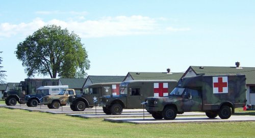 A row of military vehicles, including ambulances, parked in front of green buildings under a clear blue sky.