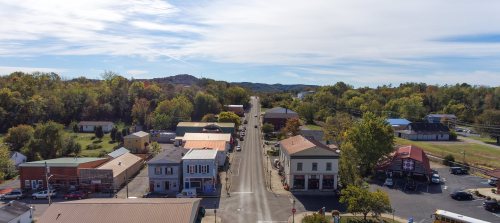 Aerial view of a small town with shops, trees, and a road lined with cars under a clear blue sky.