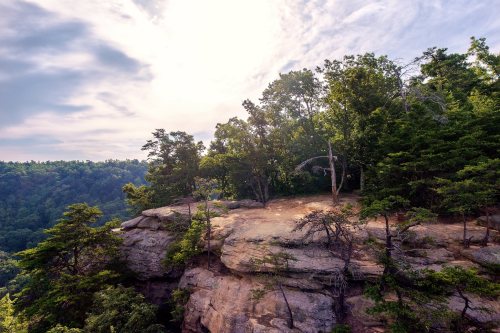 A rocky cliff surrounded by lush green trees under a cloudy sky, with a scenic view of the valley below.