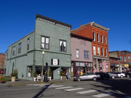 Historic buildings in a small town, featuring shops and a clear blue sky above. Cars parked along the street.