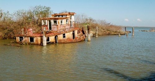 An abandoned, rusted boat partially submerged in calm water, surrounded by sparse vegetation and a clear blue sky.