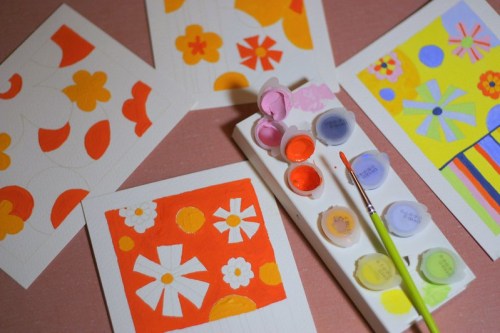 Colorful painted cards with floral designs and a palette of paints and a brush on a pink surface.
