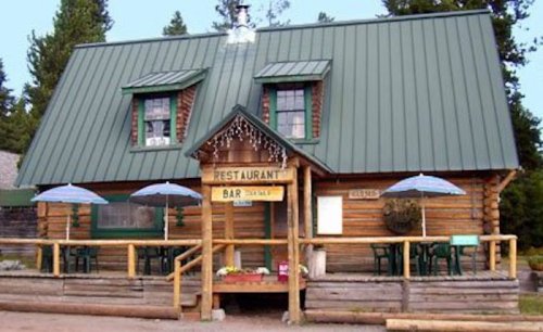 A rustic log cabin restaurant with a green metal roof, outdoor seating, and umbrellas in a forested area.