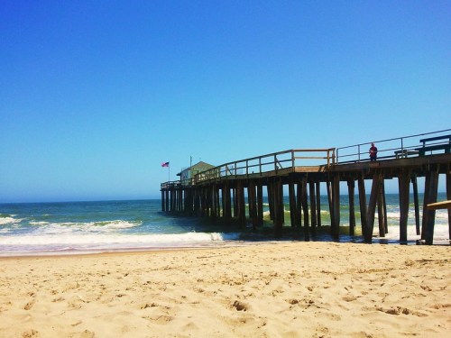 A wooden pier extends over the ocean, with gentle waves and a clear blue sky in the background.