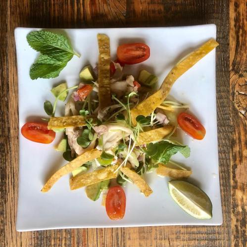 A colorful salad with avocado, cherry tomatoes, crispy tortilla strips, and fresh herbs on a white plate.