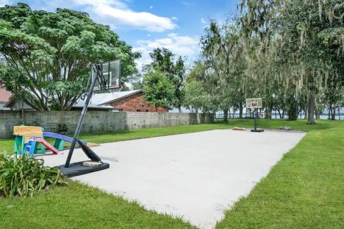 A sandy basketball court surrounded by greenery and trees, with a hoop on each end and a playset nearby.
