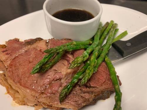 A plate with a slice of roast beef, asparagus spears, and a small bowl of sauce. A knife rests beside the meat.