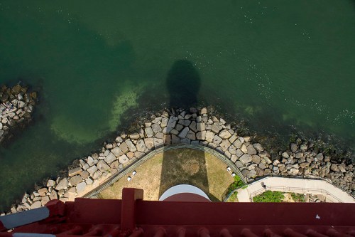 Aerial view of a rocky shoreline with a shadow cast from a nearby structure, surrounded by green water.