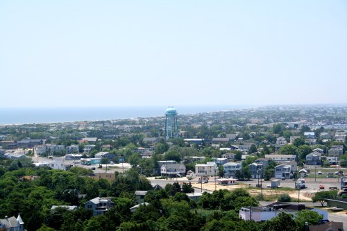 Aerial view of a coastal town with houses, greenery, and a water tower near the ocean in the background.